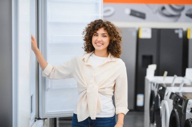 Gülümseyen bir kadın, bir mağazada çeşitli buzdolabına göz gezdirirken, mutfaktaki güncellemesi için mutlu bir şekilde karar veriyor.