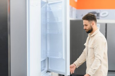 Sakallı adam yeni bir buzdolabının kapısını açarken elektronik mağazasından ev aletleri seçiyor.