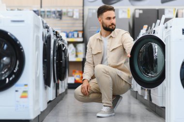 Sakallı adam bir elektronik mağazasında çamaşır makinesini kontrol ediyor, ev aletlerini seçiyor.