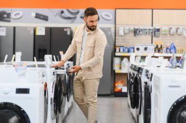 Alet mağazasından yeni çamaşır makinesi seçen, özelliklerini karşılaştıran ve satın alma kararı alan bir adam
