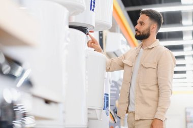 Sakallı adam ev aletleri mağazasından yeni bir su ısıtıcısı seçiyor, cihaza dokunuyor ve satın alma kararını veriyor.