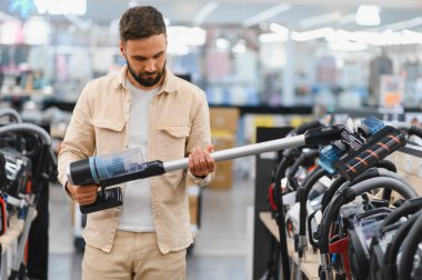 Sakallı adam yeni bir kablosuz elektrik süpürgesi seçiyor, onu elinde tutuyor, bir elektronik mağazasında.