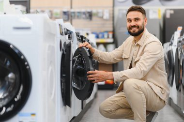 Gülümseyen müşteri, bir ev aletleri mağazasında çömelmiş çamaşır makinesinin özelliklerini inceliyor ve alışveriş deneyiminin tadını çıkarıyor.