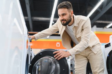 Sakallı adam ev aletleri mağazasında yeni çamaşır makinesini kontrol ediyor. Satın almadan önce ürün özelliklerini dikkatlice inceliyor.