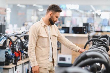 Elektronik mağazasından elektrikli süpürge seçen sakallı bir adam görüntüdeki farklı modelleri karşılaştırıyor.