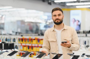 Sakallı adam yeni akıllı telefonları karşılaştırıyor, teknoloji mağazasında cep telefonunu seçiyor, tüketici elektroniği alışverişi yapıyor.