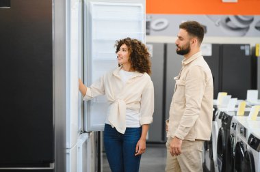 Genç çift elektronik mağazasında yeni buzdolabı seçiyor, ev aletlerini inceliyor, mutfak aletleri için alışveriş yapıyor.