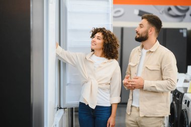 Bir çift elektronik mağazasında yeni bir buzdolabı seçiyor, seçenekleri tartışıyor ve ev ihtiyaçlarını değerlendiriyor.
