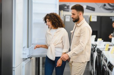 Çift, çeşitli ev aletleri arasından yeni bir buzdolabı seçmeye odaklanarak bir elektronik mağazasında seçenekleri araştırıyor.