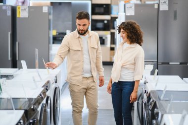 Ev aletleri mağazasında çeşitli çamaşır makinelerini karşılaştıran çift, ev ihtiyaçlarının özelliklerini ve seçeneklerini değerlendiriyor.