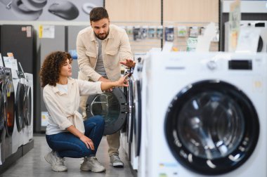 Bir çift, bir mağazadaki çamaşır makinelerini karıştırıyor, seçenekleri tartışıyor ve evleri için markaları karşılaştırıyor.