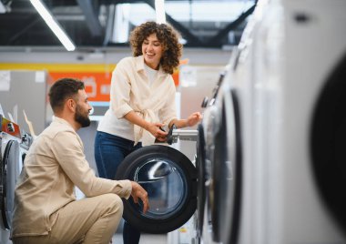 Çift bir elektronik mağazasında çamaşır makinelerini hevesle inceliyor, özellikleri tartışıyor ve satın alma kararı veriyor