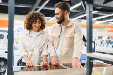 Mutlu çift, bir ev aletleri mağazasında alışveriş yaparken indüksiyon fırınını test ediyor ve mutfak malzemelerini birlikte seçmenin tadını çıkarıyor.