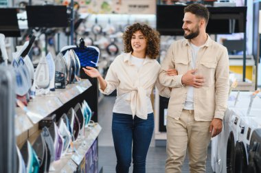 Genç çift elektronik mağazasında yeni ev aletleri seçiyor. Kadın farklı ütü modelleri gösteriyor.