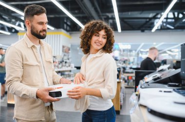 Robotik süpürge tutan genç bir çift, elektronik mağazasında birlikte alışveriş yapıyorlar, akıllı evleri için satın alma kararı veriyorlar.