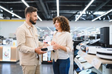Mutlu çift, elektrikli süpürge tutarken gülümsüyor, canlı bir elektronik mağazasında çeşitli ev aletlerini karıştırıyor.