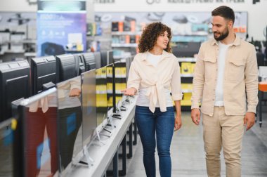 Çift bir elektronik mağazasından televizyon seçiyor, ev aletlerini incelerken ve satın aldıklarını düşünürken seçenekleri tartışıyor.