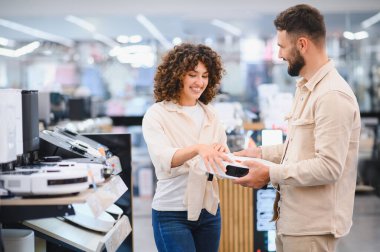 Gülümseyen bir çift elektronik mağazasından yeni bir robot elektrikli süpürge seçiyor, ev aletleri alışverişi yapıyor.