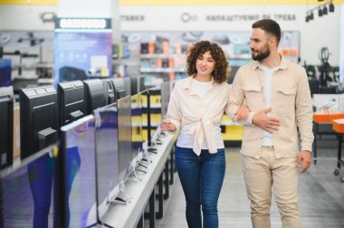 Bir tüketici elektronik mağazasında kol kola yürüyen mutlu çift, ev eğlenceleri için yeni bir televizyon seçiyorlar.