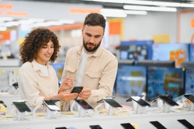 Genç çift çeşitli akıllı telefonları karşılaştırırken tüketici elektroniği mağazasındaki en iyi seçeneği seçiyor, özellikleri ve fiyatları değerlendiriyor