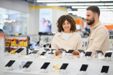 Çift, modern bir perakende piyasasının ortasındaki yeni satın alma seçeneklerini göz önünde bulundurarak bir elektronik mağazasında çeşitli akıllı telefonları inceliyor.