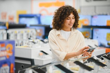 Genç bir kadın telefonunu kullanıyor ve akıllı telefonları karşılaştırıyor. Elektronik mağazasından yeni bir cep telefonu seçip alıyor.