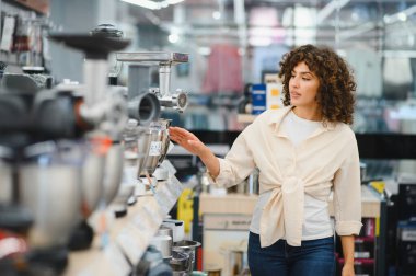 Bir perakende mağazasında ev aletlerine bakarken mutfak robotunu işaret eden genç bir kadın, evi için dikkatlice ürünleri seçiyor.