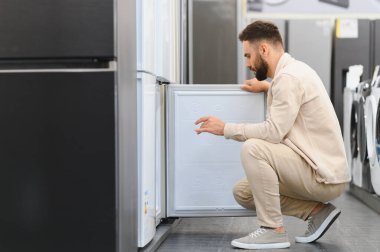 Sakallı adam, çömelmiş ve buzdolabının kapısını açıyor. Elektronik mağazasından ev aletlerini seçiyor.