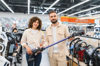 Mutlu çift yeni bir kablosuz elektrik süpürgesi seçiyor, onu ellerinde tutuyorlar, büyük bir elektronik mağazasında birlikte alışveriş yapıyorlar.