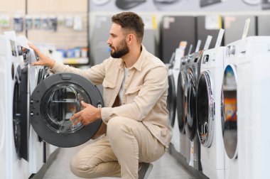 Modern bir elektronik mağazasında ev aletleri seçerken bir çamaşır makinesinin özelliklerini inceleyen sakallı bir adam.
