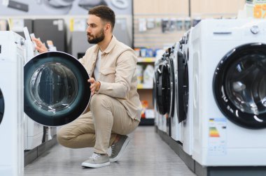 Sakallı adam çömelmiş, mağazadan yeni çamaşır makinesi seçiyor, satın almadan önce özelliklerini inceliyor.
