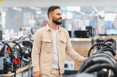 Gülümseyen adam yeni bir elektrikli süpürge seçiyor. Bir ev aletleri mağazasının koridorlarında geziniyor, alışveriş deneyiminin tadını çıkarıyor.