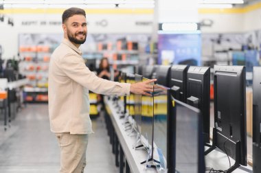 Tüketici elektronik mağazasından modern bir televizyon seçen sakallı bir adam, vitrindeki farklı modelleri karşılaştırıyor.