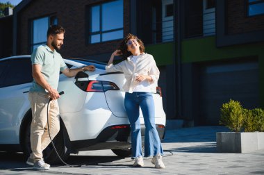 Modern evlerinin önünde elektrikli araçlarını şarj eden mutlu ev sahipleri, sürdürülebilir ulaşım ve yeşil enerji çözümlerini kucaklıyorlar.