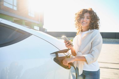 Mutlu kadın elektrik kablosunu elektrikli araca bağlıyor ve evinde akıllı telefon uygulaması kullanıyor.