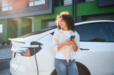 Modern evin dışında elektrikli araba şarj ederken akıllı telefon kullanan gülümseyen genç kadın, yenilenebilir enerji ve çevre dostu yaşam tarzını ön plana çıkarıyor