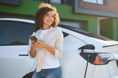 Elektrikli arabası modern evinin önünde şarj olurken elinde akıllı telefon tutan mutlu kadın sürdürülebilir ve yeşil yaşam tarzının tadını çıkarıyor.