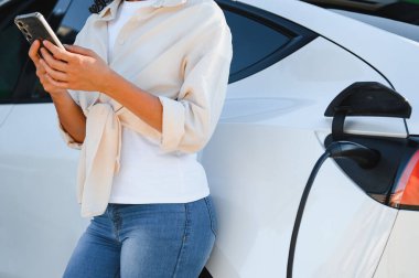 Elektrikli arabası evinde şarj olurken akıllı telefonunu kullanan genç bir kadın modern ve sürdürülebilir bir yaşam tarzı sergiliyor.