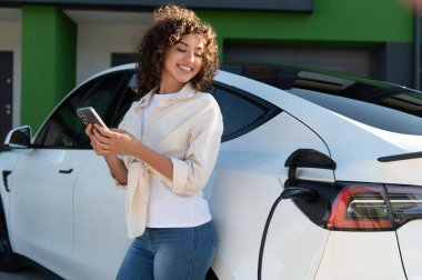 Gülümseyen genç bir kadın elektrikli arabasını şarj ediyor, yenilenebilir enerjiyi sembolize ediyor ve modern yerleşim bölgesinde sürdürülebilir yaşamı simgeliyor.