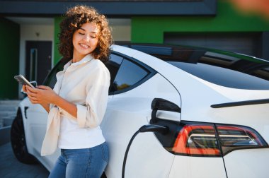 Evde elektrikli arabasını şarj ederken cep telefonu kullanan genç bir kadın, sürdürülebilir ulaşım ve yeşil enerji çözümlerini kucaklıyor