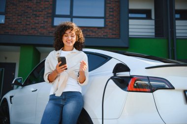 Genç bir kadın elektrikli arabası modern evinin önünde şarj olurken akıllı telefonunu kullanıyor, sürdürülebilir ulaşımı kucaklıyor.