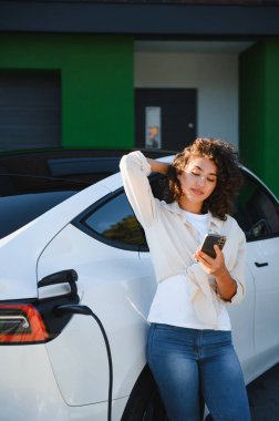 Evde elektrikli araba şarj ederken akıllı telefon kullanan genç bir kadın sürdürülebilir ve uygun bir yaşam tarzı sergiliyor.