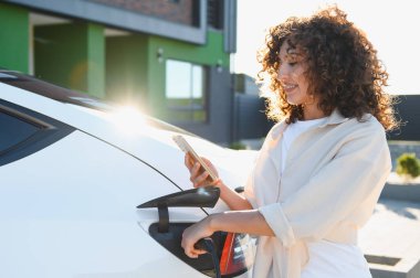 Genç bir kadın evinin yakınında elektrikli arabasını şarj ediyor ve süreci izlemek için akıllı telefon uygulaması kullanıyor.