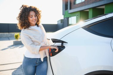 Gülümseyen genç kadın elektrikli arabayı modern evinin dışına bağlıyor, sürdürülebilir yaşam ve yenilenebilir enerji uygulamalarını vurguluyor