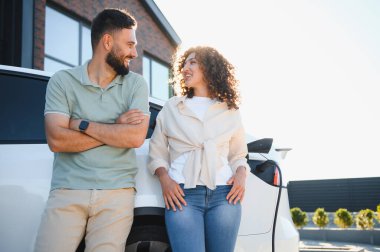 Genç çift modern evlerinde elektrikli araçlarını şarj ederken gülümsüyor, sürdürülebilir yaşam ve ekolojik bilinçli tercihler sergiliyor