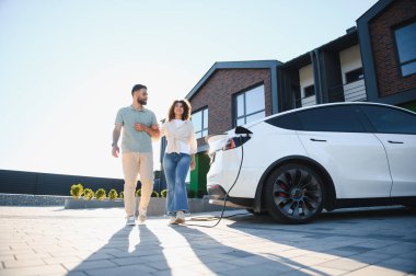 Elektrikli arabaları şarj olurken, sürdürülebilir ve yeşil yaşam tarzlarını kucaklarken, modern evlerinin yanında yürüyen mutlu çift.