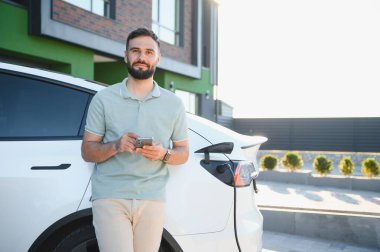 Modern evinde elektrikli araba şarj ederken akıllı telefon kullanan ilerici bir adam.