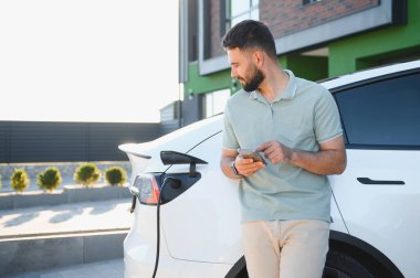 İş adamı evde elektrikli arabasını şarj ederken telefonu kullanıyor.
