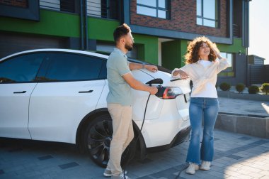 Genç çift modern evlerinde elektrikli araçlarını şarj ediyor, çevre dostu ulaşım ve yenilenebilir enerji çözümlerine bağlılıklarını sergiliyorlar.