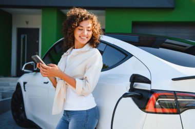 Modern evinin önünde elektrikli araba şarj ederken akıllı telefon kullanan gülümseyen genç kadın yenilenebilir enerjiyi ve sürdürülebilir yaşamı sembolize ediyor.